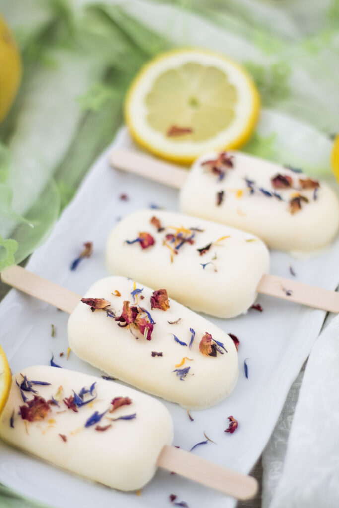 Veganes Zitronen-Mango-Eis am Stil mit bunten Blüten und Zitronenscheiben